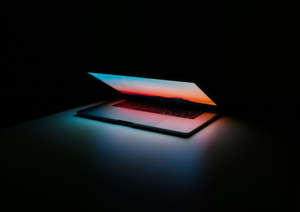 A partially open laptop, on a dark surface and set amongst a black background. The only visible light is emanating from the partially-open laptop screen.